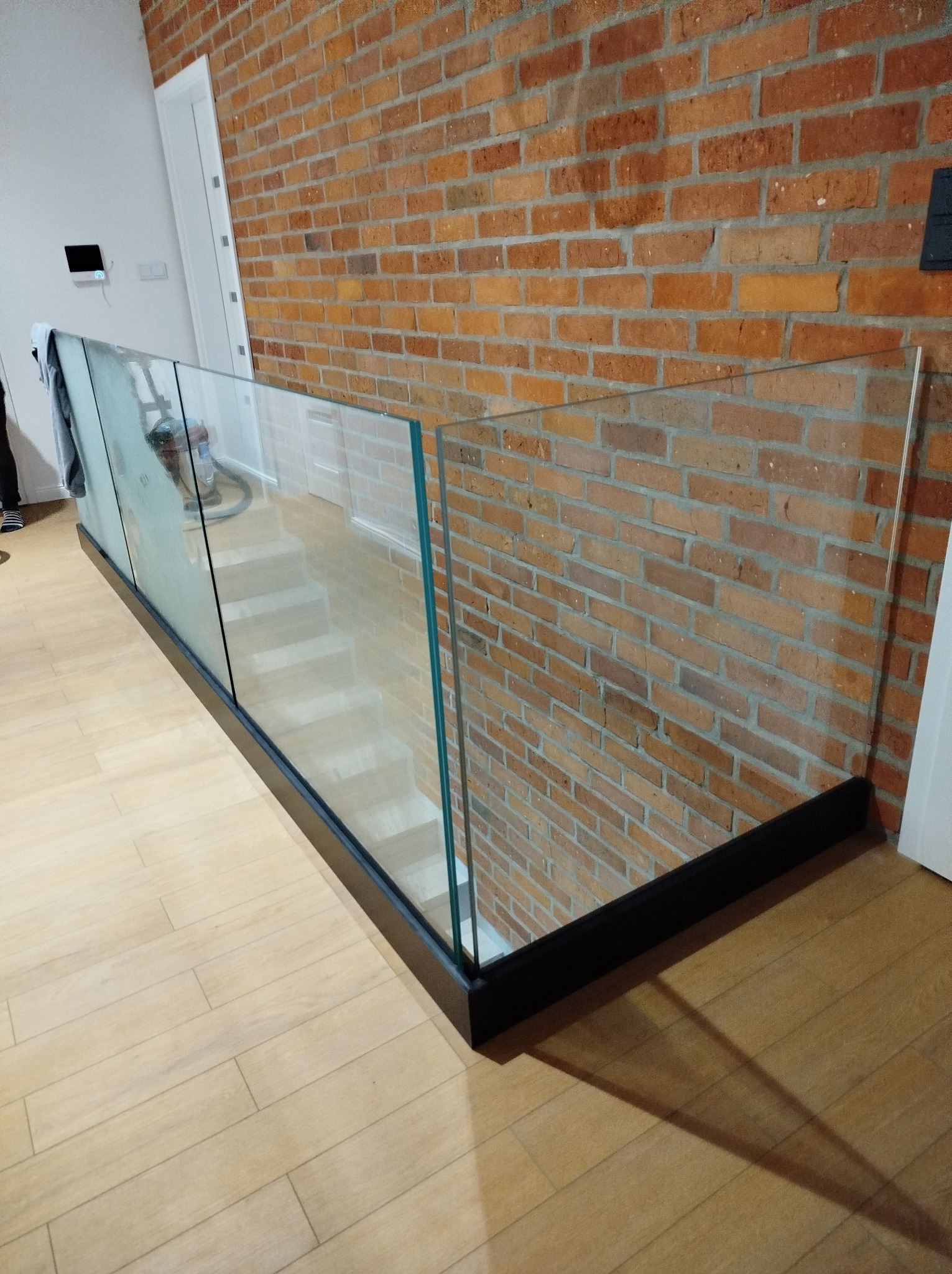 Szklana balustrada z czarną ramą w nowoczesnym wnętrzu z ceglaną ścianą i drewnianą podłogą. Widok z góry na schody. Minimalistyczny design.