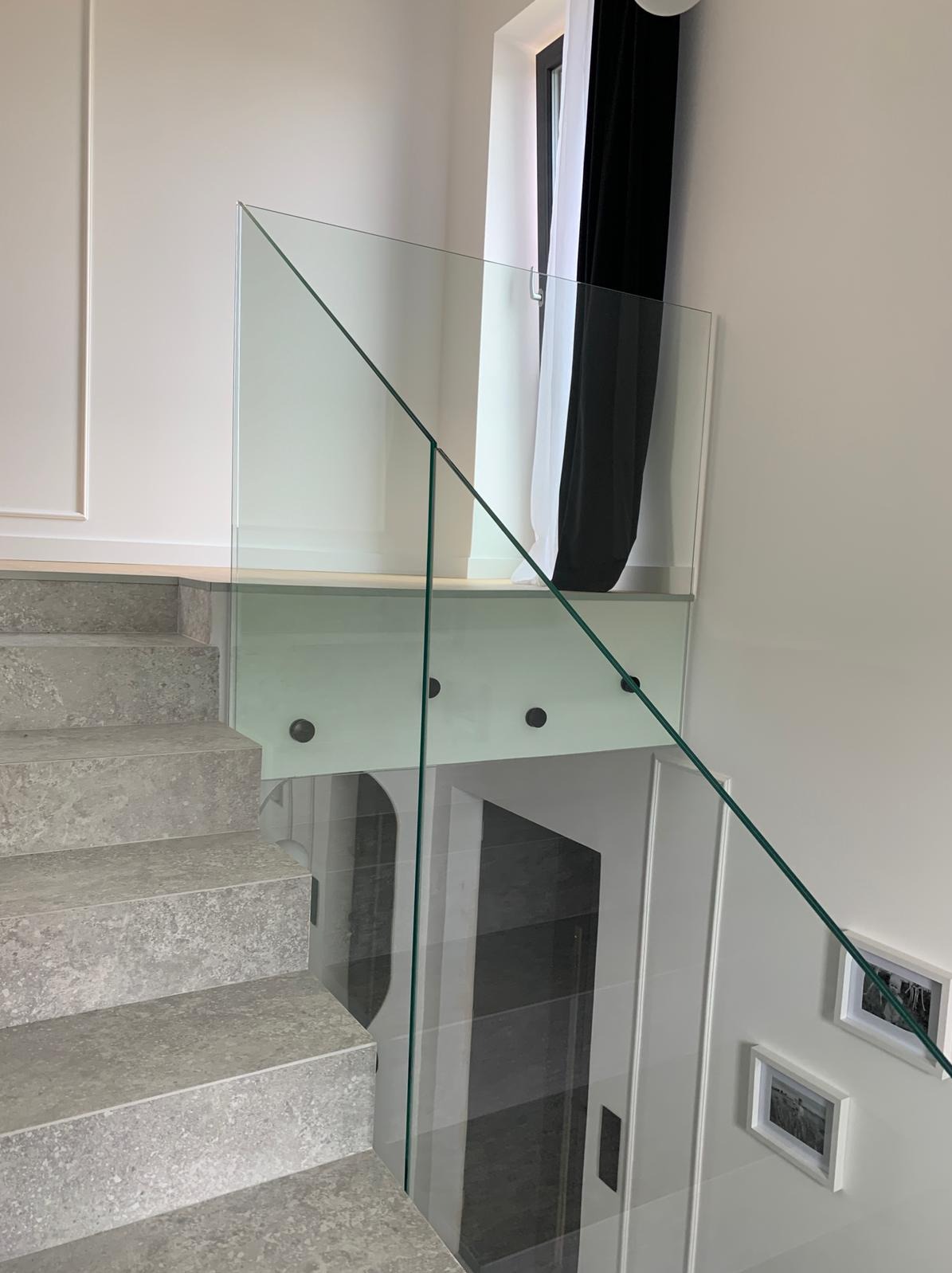 Szklana balustrada z mocowaniami punktowymi przy schodach z szarego kamienia. W tle okno z czarno-białą zasłoną i dekoracyjne ramki na ścianie.