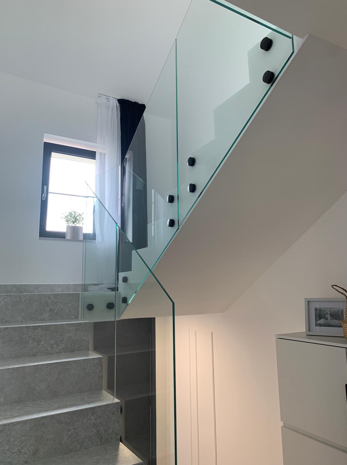 Nowoczesna, szklana balustrada schodowa z czarnymi mocowaniami. Widok z dołu, jasne wnętrze z oknem i zasłonami. Minimalistyczny design.