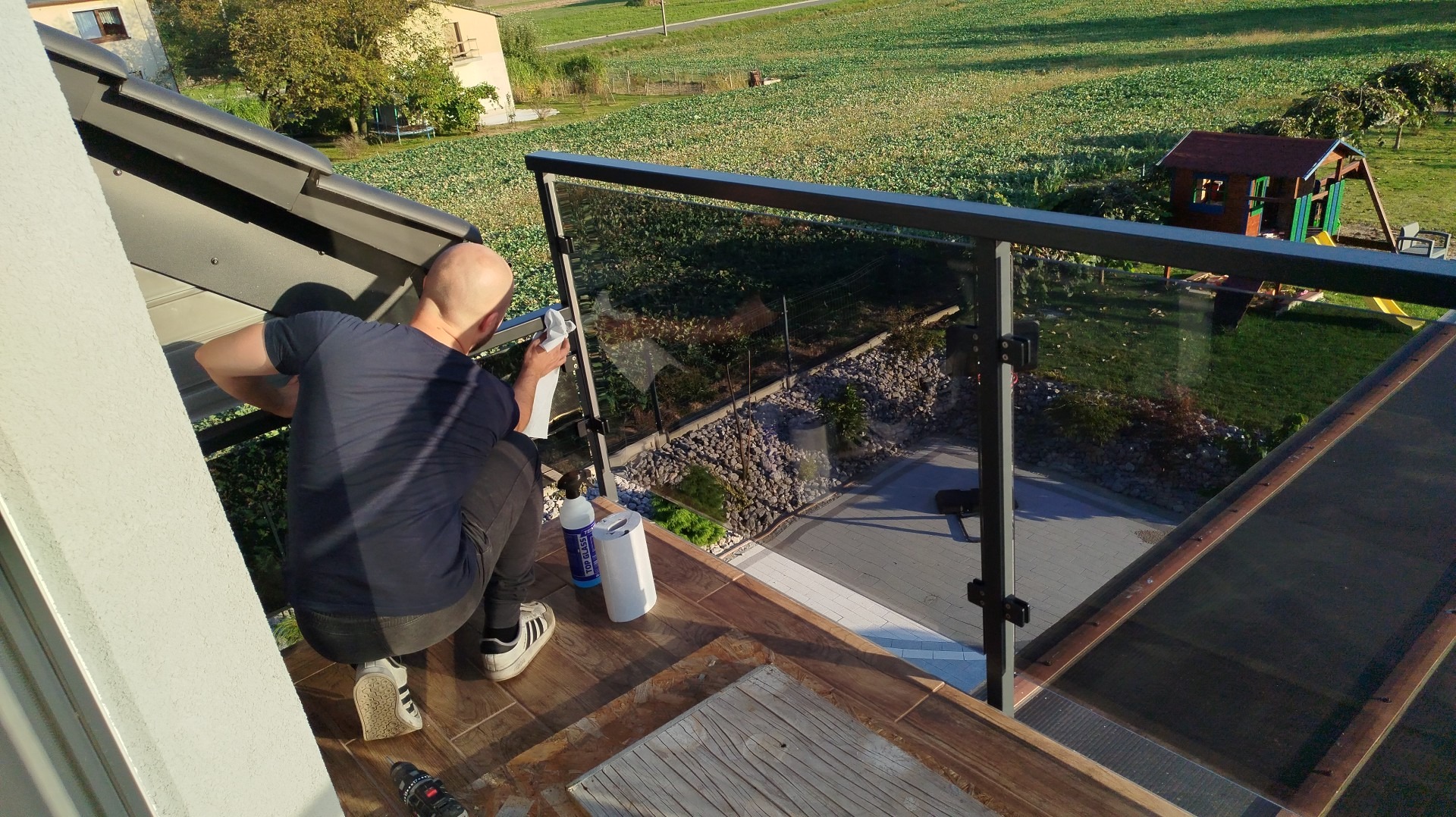 Osoba czyści szklaną balustradę na tarasie z widokiem na ogród. Widoczne środki czystości i narzędzia. Ujęcie z góry, w słoneczny dzień.