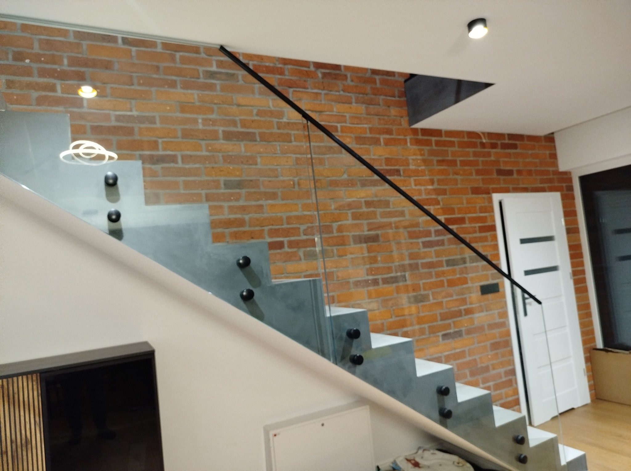 Szklana balustrada schodowa z czarnymi uchwytami na tle ceglanej ściany i białych drzwi. Minimalistyczny design, nowoczesne wykończenie.