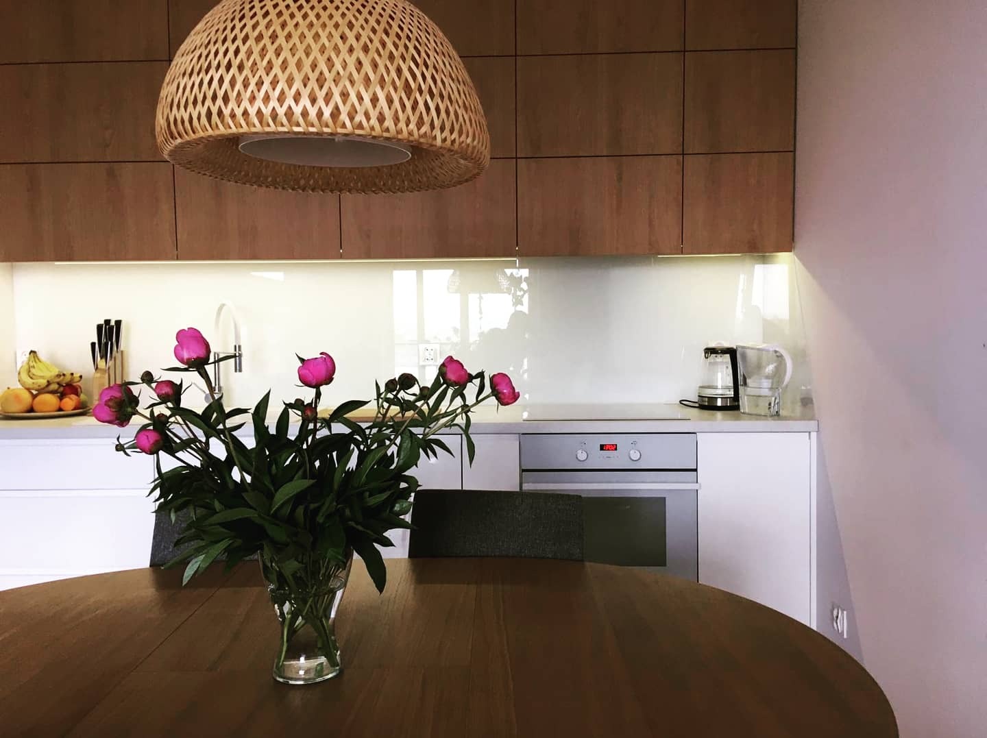 Nowoczesna kuchnia z białymi frontami i drewnianymi szafkami wiszącymi, ozdobiona szklanym panelem nad blatem i bukietem różowych piwonii na okrągłym stole.