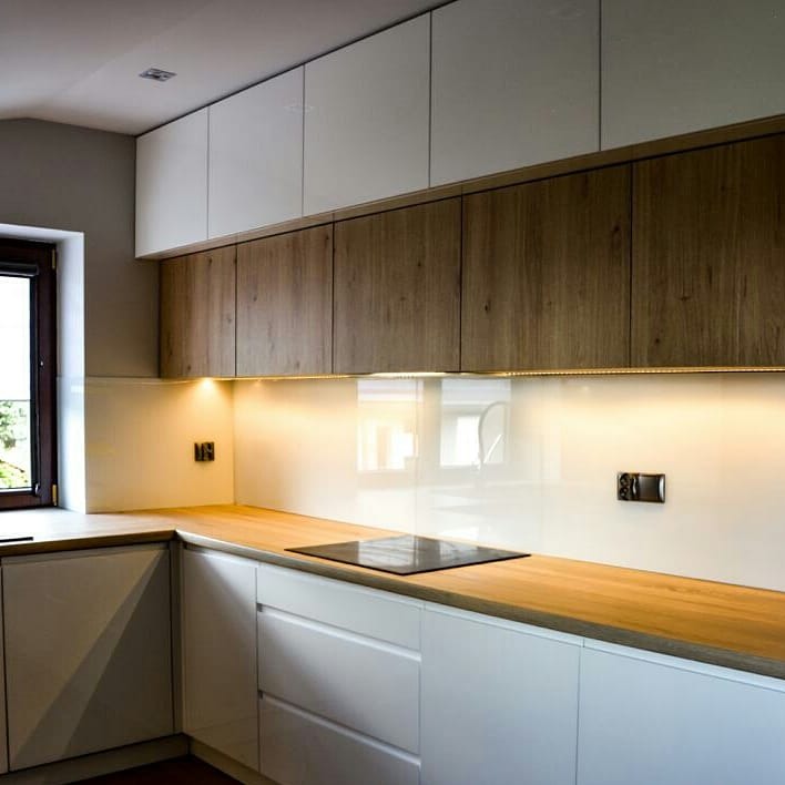 Nowoczesna kuchnia z białymi szafkami, drewnianymi blatami i szklanym panelem nad blatem, podświetlonym listwą LED.