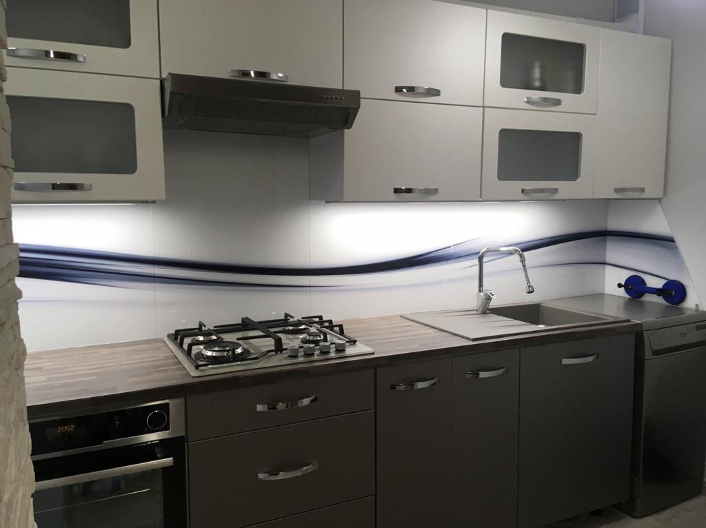 Kuchnia z szarą zabudową i szklanym panelem ściennym z motywem fali. Na blacie kuchenka gazowa i zlew. Widoczne przyssawki do szkła.