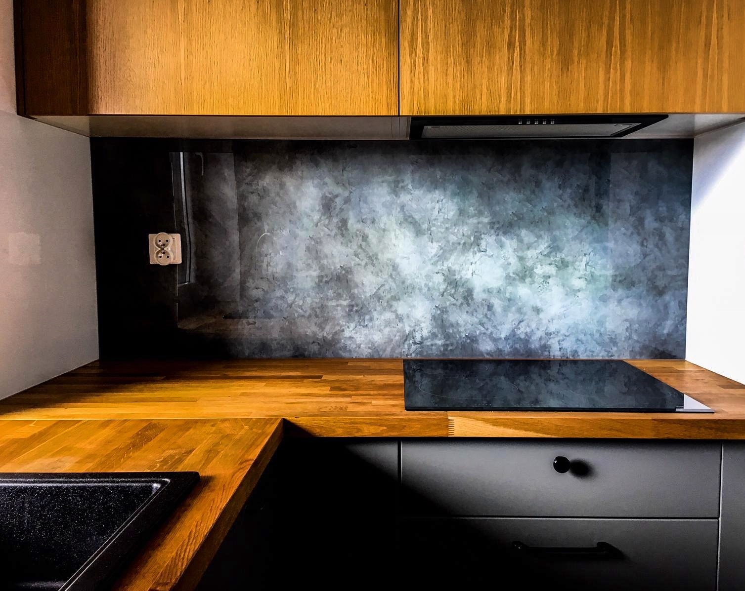 Szklany panel kuchenny z abstrakcyjnym wzorem w odcieniach szarości, zamontowany między drewnianym blatem a szafkami. Widoczne gniazdko elektryczne i płyta indukcyjna.