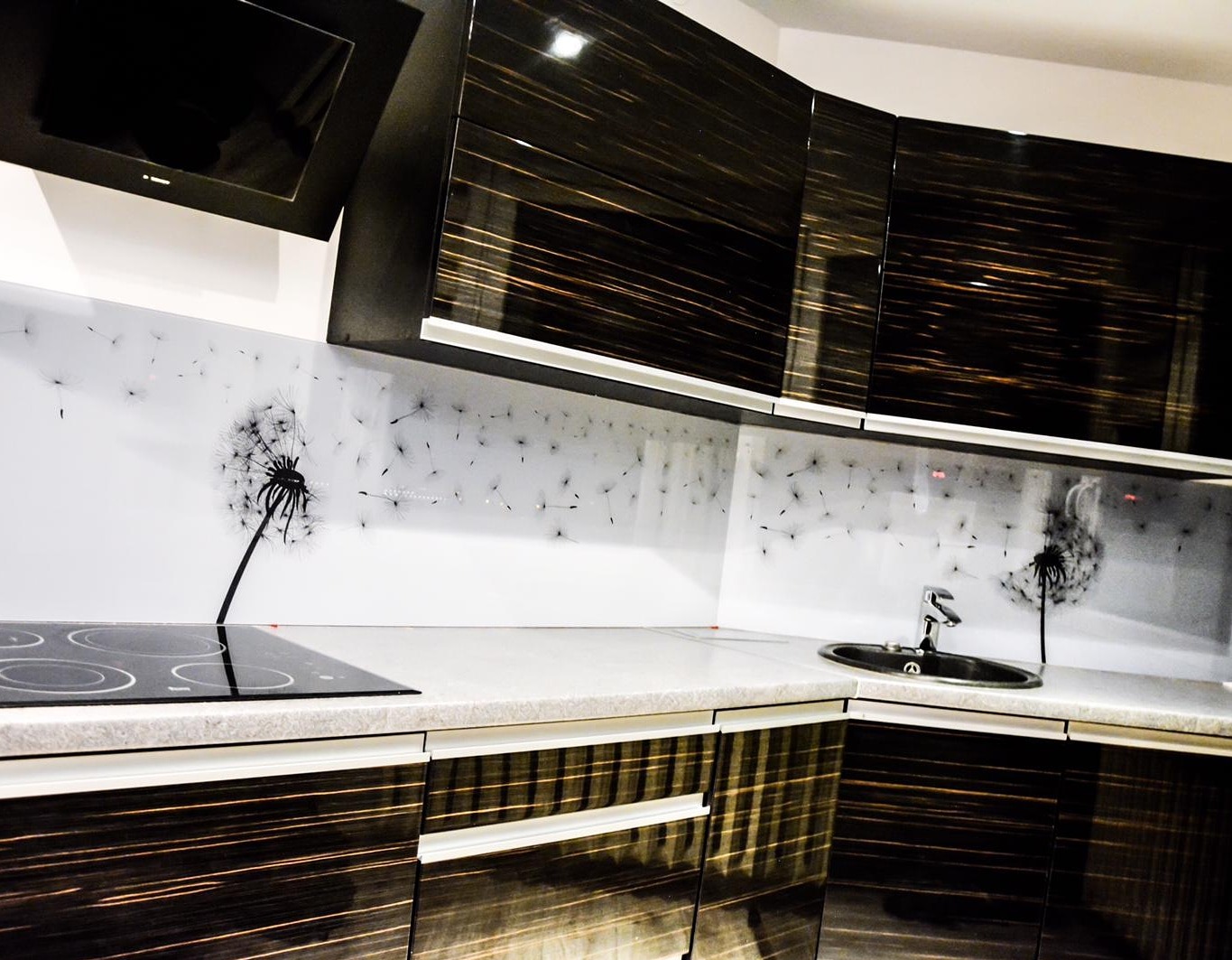 Nowoczesna kuchnia z czarnymi szafkami i szklanym panelem ściennym z motywem dmuchawca. Bateria umywalkowa i płyta indukcyjna widoczne na blacie.