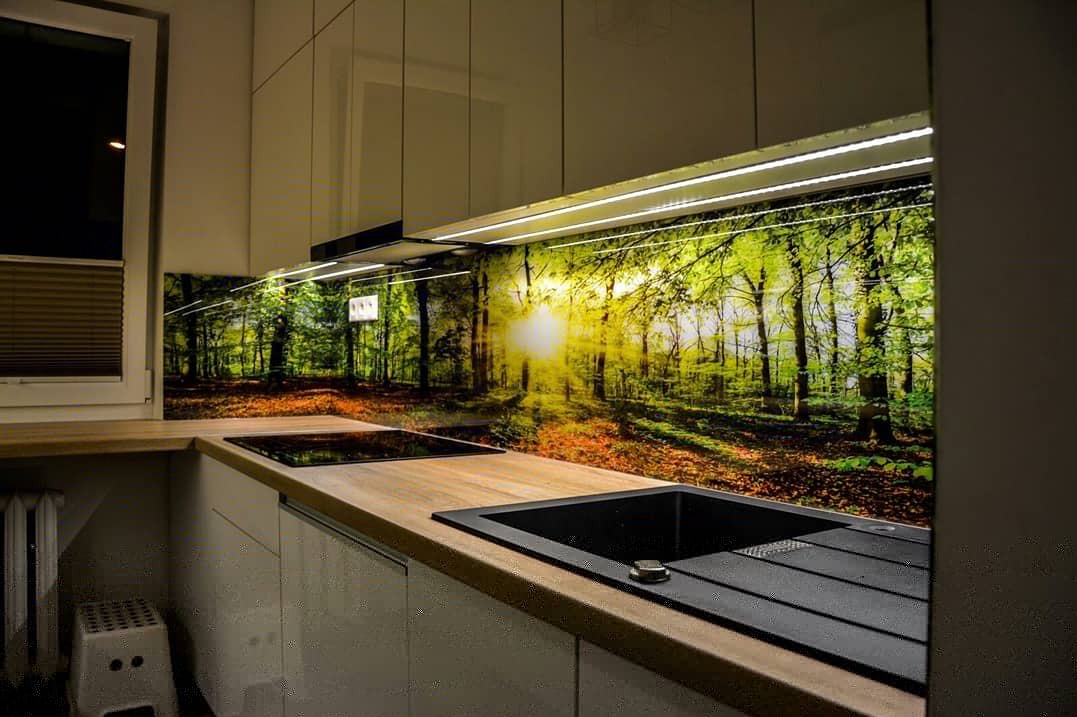 Szklany panel kuchenny z motywem leśnym podświetlony LED, zabudowa meblowa w odcieniach bieli, czarny zlew i płyta grzewcza. Nowoczesny design kuchni.