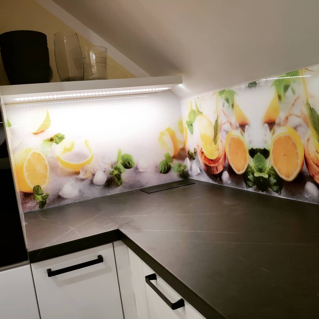 Szklany panel kuchenny z motywem cytryn i mięty, podświetlony LED, zamontowany nad blatem z czarnego kamienia i białymi szafkami z czarnymi uchwytami.