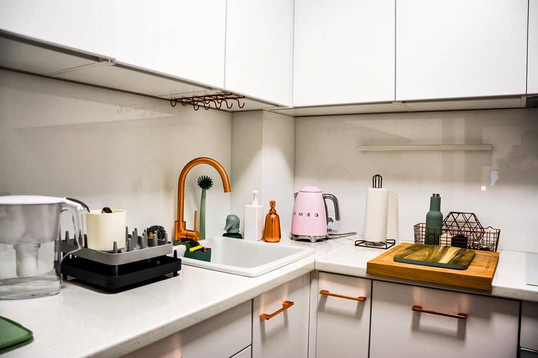 Nowoczesna kuchnia z białymi szafkami, złotą armaturą i szklanym panelem nad blatem. Widoczny zlewozmywak, suszarka do naczyń i akcesoria kuchenne.