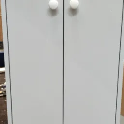 Szara szafka z dwiema białymi, okrągłymi gałkami na tle warsztatu. Prosty, minimalistyczny design. Widoczne elementy konstrukcyjne i warsztatowe w tle.