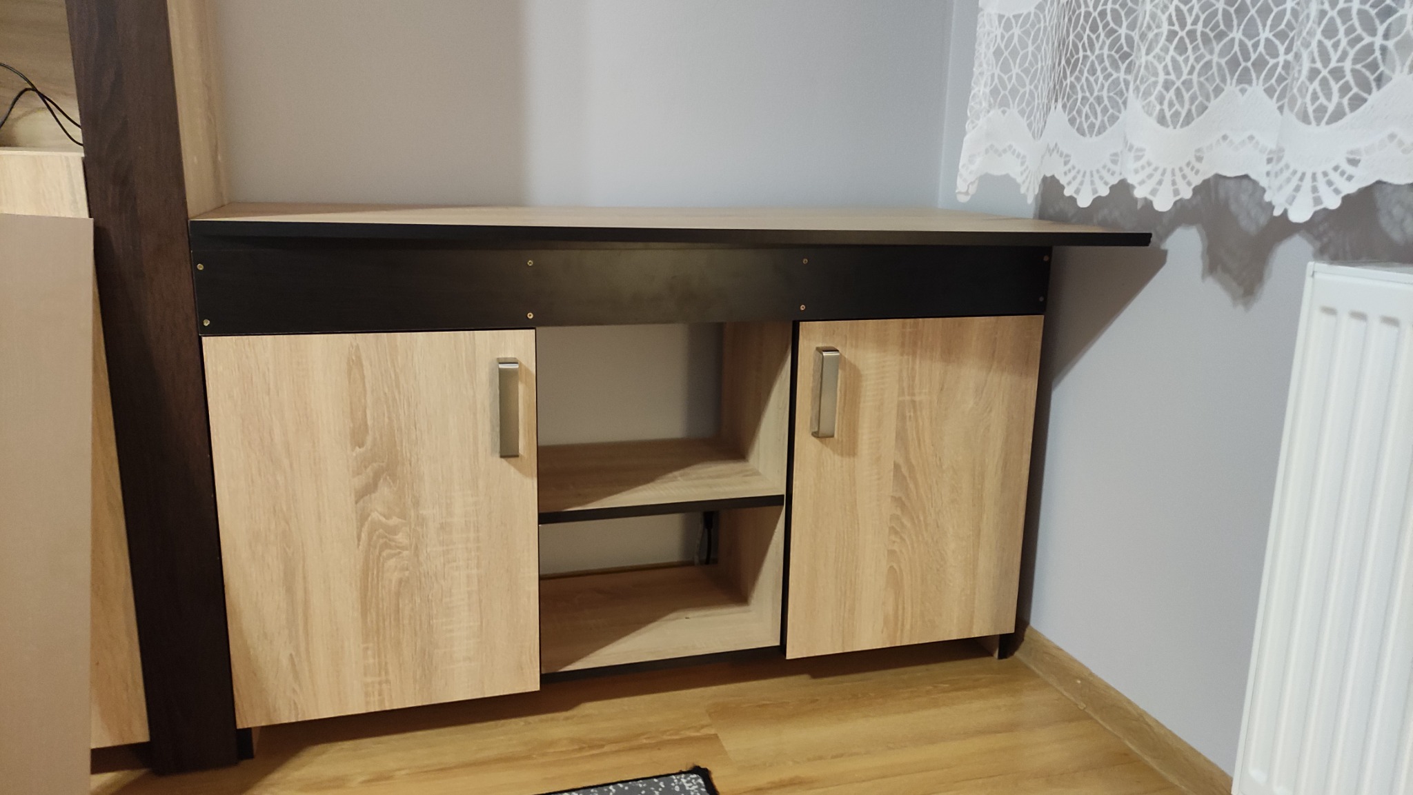 Drewniana szafka z czarnym blatem i metalowymi uchwytami, widoczna w Radomiu. Szafka posiada półki w otwartej części i zamknięte szafki z drzwiczkami.