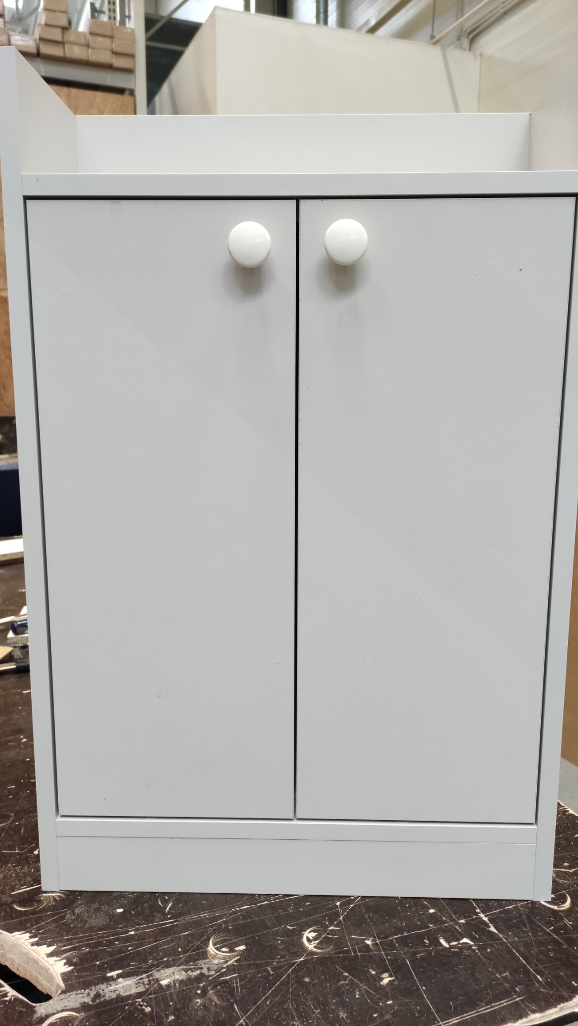 Szara szafka z dwiema białymi, okrągłymi gałkami na tle warsztatu. Prosty, minimalistyczny design. Widoczne elementy konstrukcyjne i warsztatowe w tle.