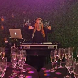 Patrycja Neodym - DJ kobieta za konsoletą, uśmiechnięta, z gestem 'V' dłońmi. W tle zielona ściana z napisem 'Let's party!', stoliki z kieliszkami, fioletowe oświetlenie.