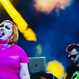 Patrycja Neodym - Ekstrawagancka wokalistka w różowej sukience śpiewa do mikrofonu na scenie, w tle muzyk z laptopem i oświetlenie sceniczne.