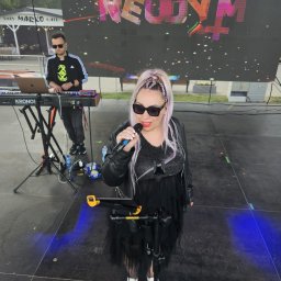 Patrycja Neodym - Zespół muzyczny na scenie: wokalistka z mikrofonem i klawiszowiec grający na keyboardzie Kronos, na telebimie logo zespołu 'Neodym', plenerowa scena.