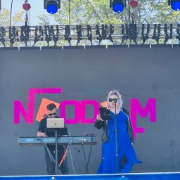 Patrycja Neodym - Zespół muzyczny na scenie plenerowej: wokalistka w niebieskiej sukience i klawiszowiec z laptopem grają na syntezatorze Korg Kronos, oświetlenie sceniczne.