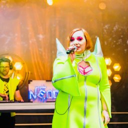 Patrycja Neodym - Wokalistka w neonowej kreacji śpiewa do mikrofonu na scenie. W tle DJ w słuchawkach i neonowy napis 'NEON'.