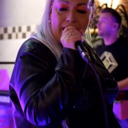 Patrycja Neodym - Wokalistka z mikrofonem, w skórzanej kurtce, ze srebrnymi włosami, śpiewa na scenie oświetlonej fioletowym światłem. W tle drugi muzyk.