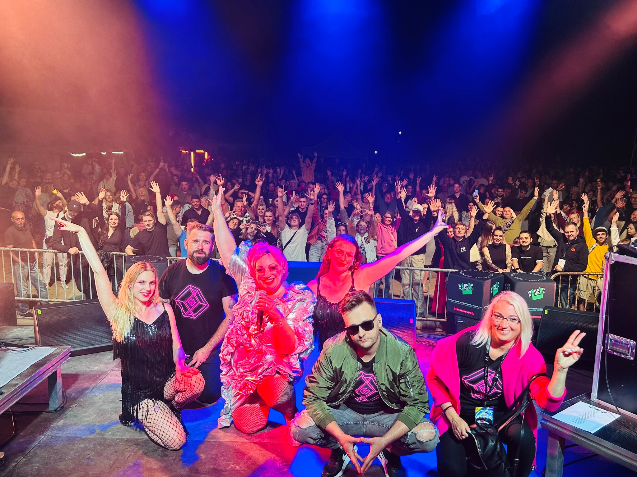 Zespół muzyczny na scenie podczas koncertu na tle rozentuzjazmowanego tłumu, oświetlony czerwonym i niebieskim światłem, członkowie zespołu w charakterystycznych strojach z logo na koszulkach.