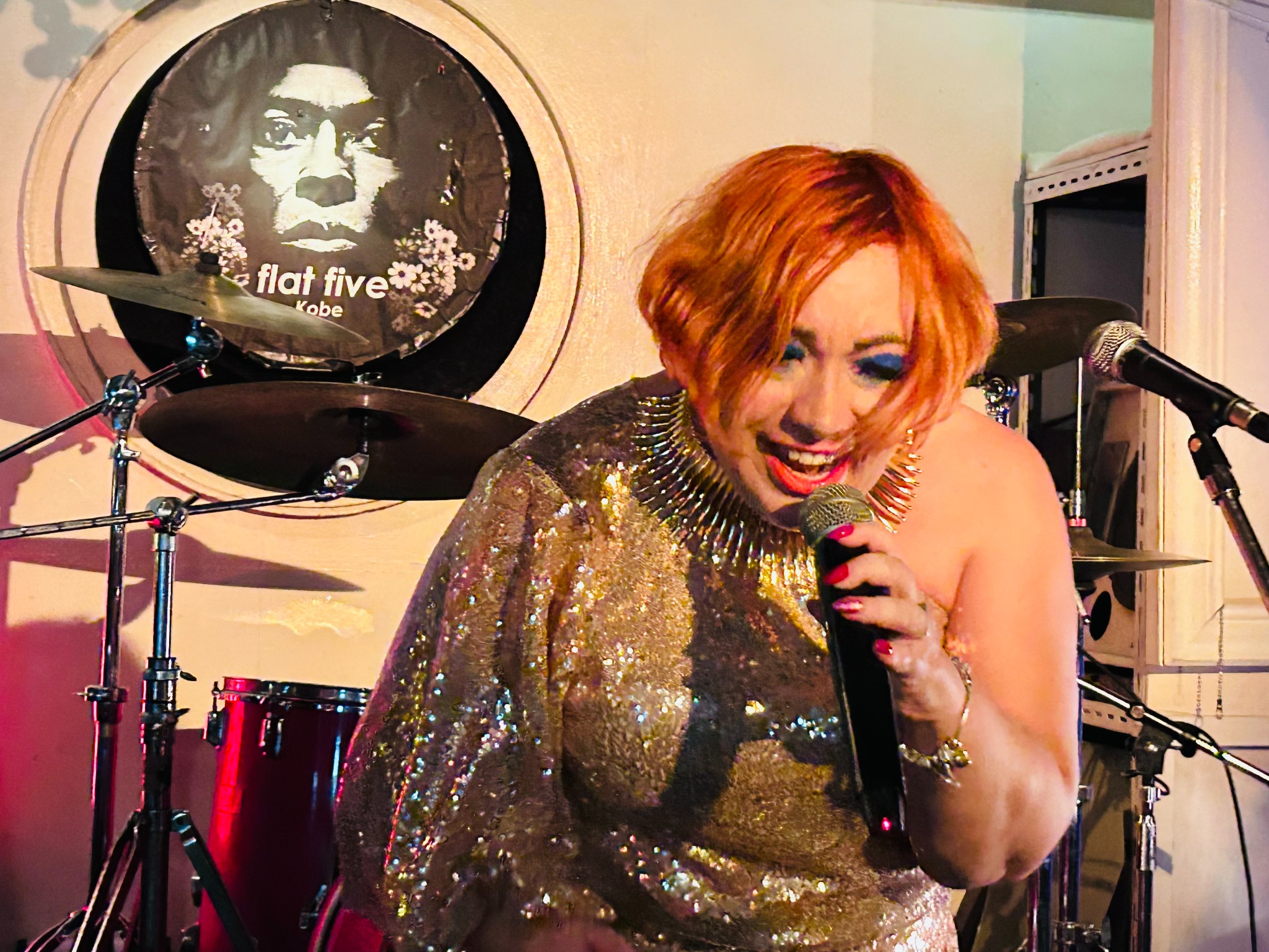 Wokalistka z krótkimi, rudymi włosami śpiewa do mikrofonu w błyszczącej sukni, na tle perkusji z plakatem 'Flat Five Kobe'.