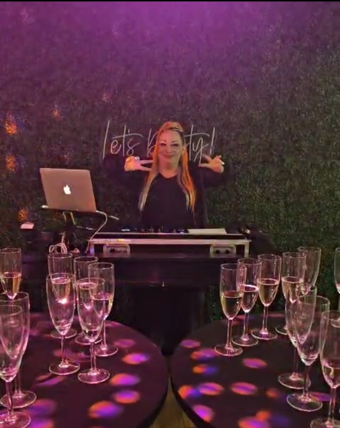 DJ kobieta za konsoletą, uśmiechnięta, z gestem 'V' dłońmi. W tle zielona ściana z napisem 'Let's party!', stoliki z kieliszkami, fioletowe oświetlenie.