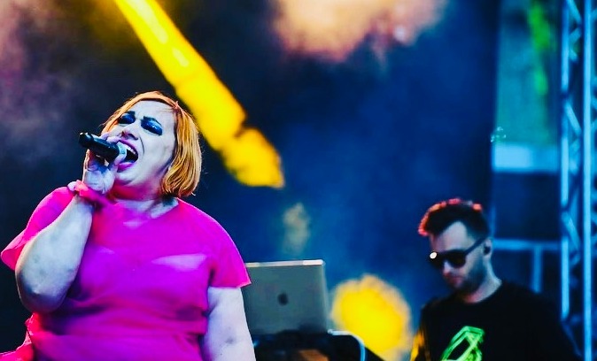 Ekstrawagancka wokalistka w różowej sukience śpiewa do mikrofonu na scenie, w tle muzyk z laptopem i oświetlenie sceniczne.