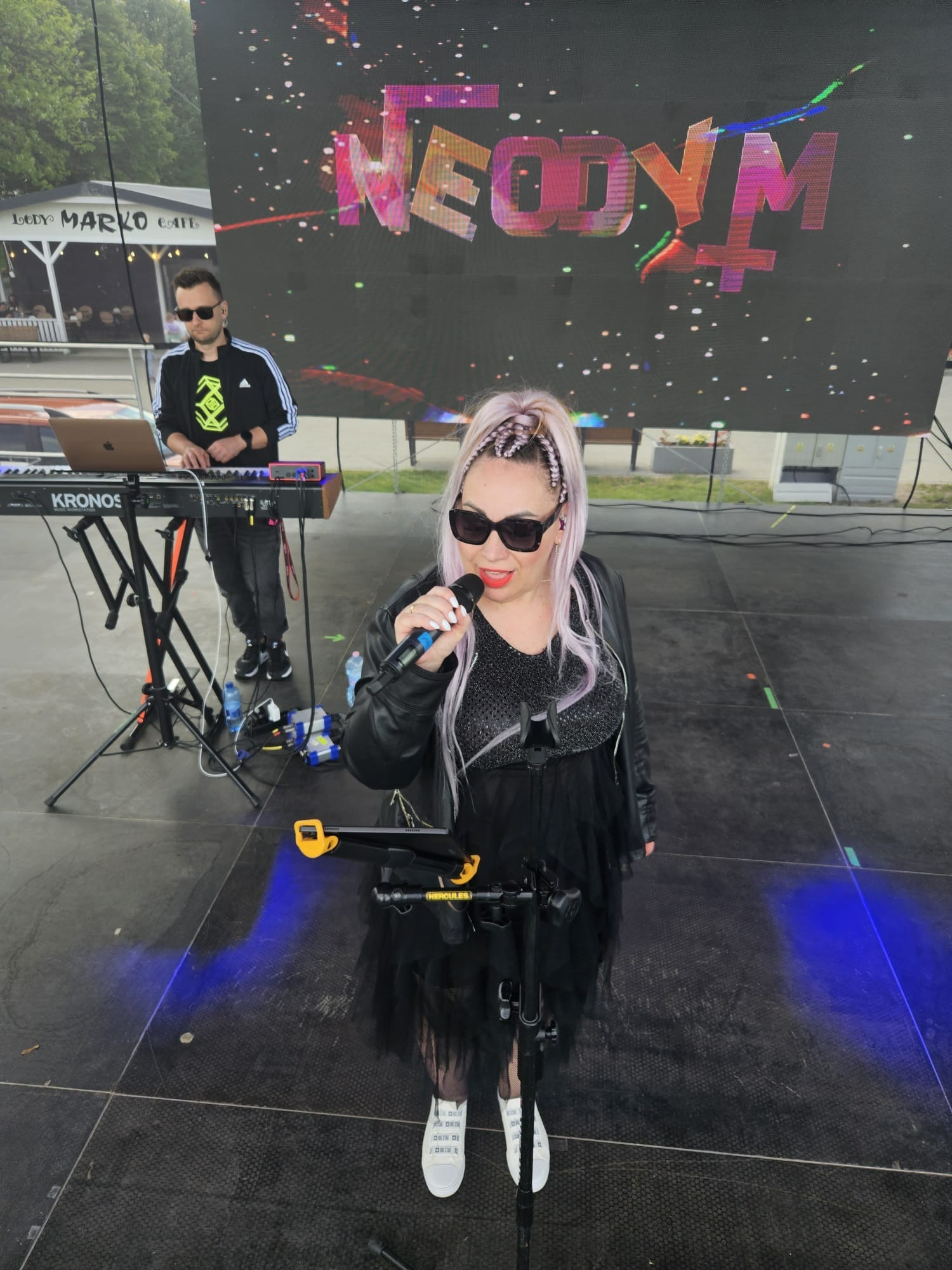 Zespół muzyczny na scenie: wokalistka z mikrofonem i klawiszowiec grający na keyboardzie Kronos, na telebimie logo zespołu 'Neodym', plenerowa scena.