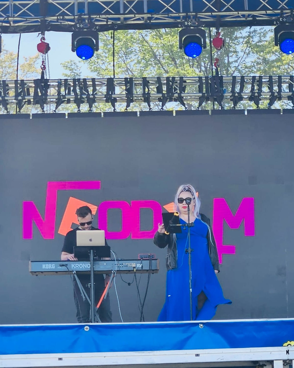 Zespół muzyczny na scenie plenerowej: wokalistka w niebieskiej sukience i klawiszowiec z laptopem grają na syntezatorze Korg Kronos, oświetlenie sceniczne.