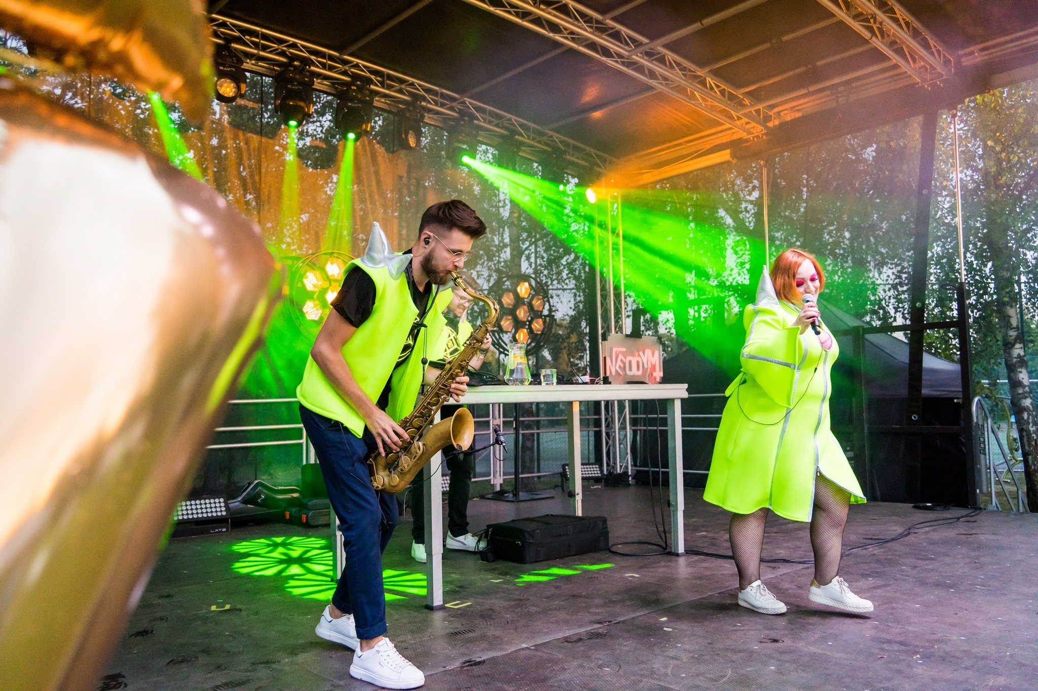 Występ zespołu na scenie: saksofonista w neonowej kamizelce i wokalistka w neonowej sukience, podświetlenie zielonymi reflektorami, nietypowe stroje.