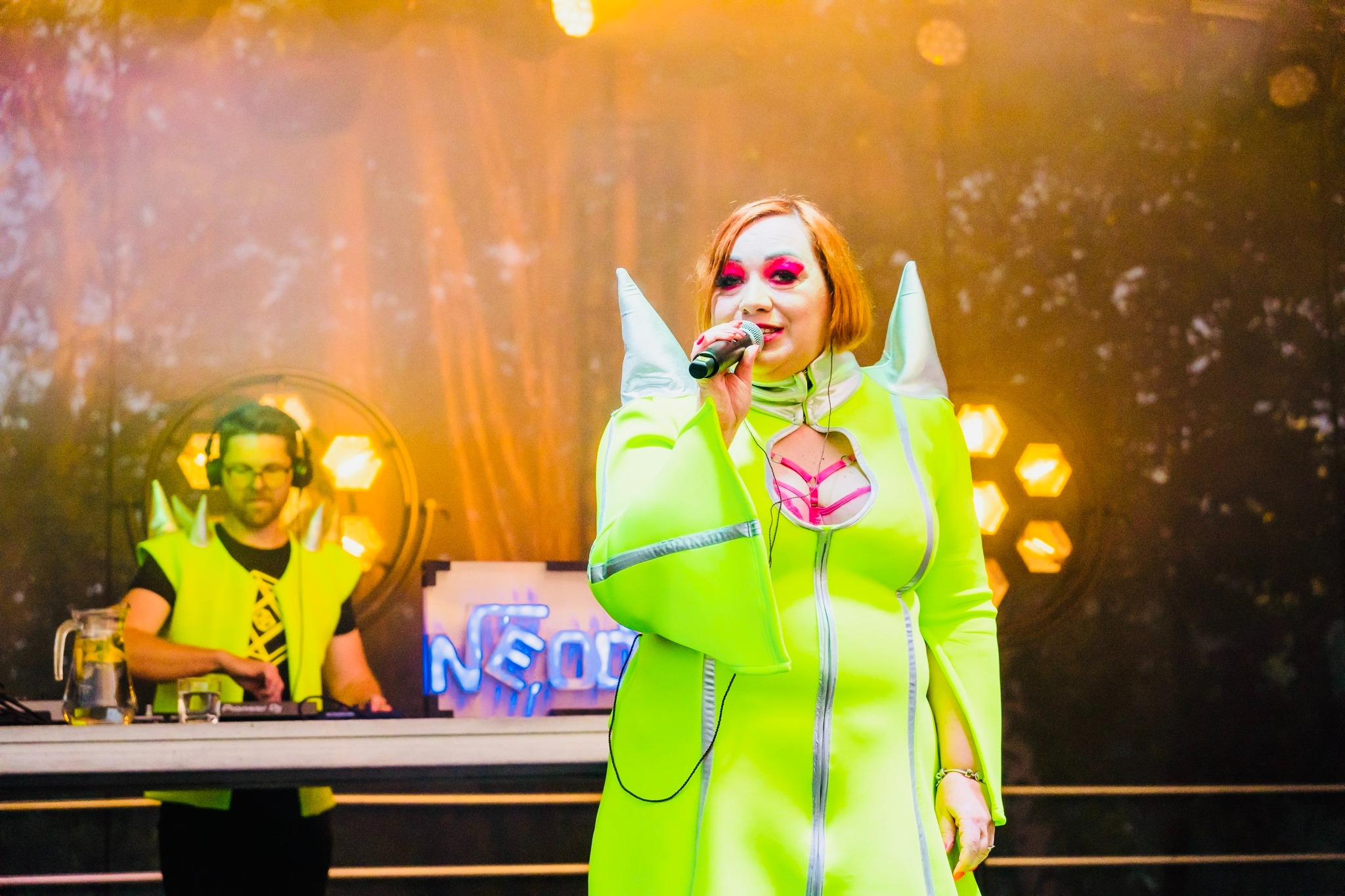 Wokalistka w neonowej kreacji śpiewa do mikrofonu na scenie. W tle DJ w słuchawkach i neonowy napis 'NEON'.