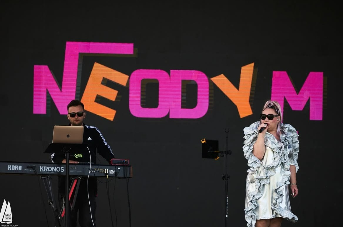 Występ zespołu Neodym: kobieta w srebrnej sukience śpiewa do mikrofonu, obok mężczyzna gra na keyboardzie Korg Kronos z laptopem na tle neonowego logo.