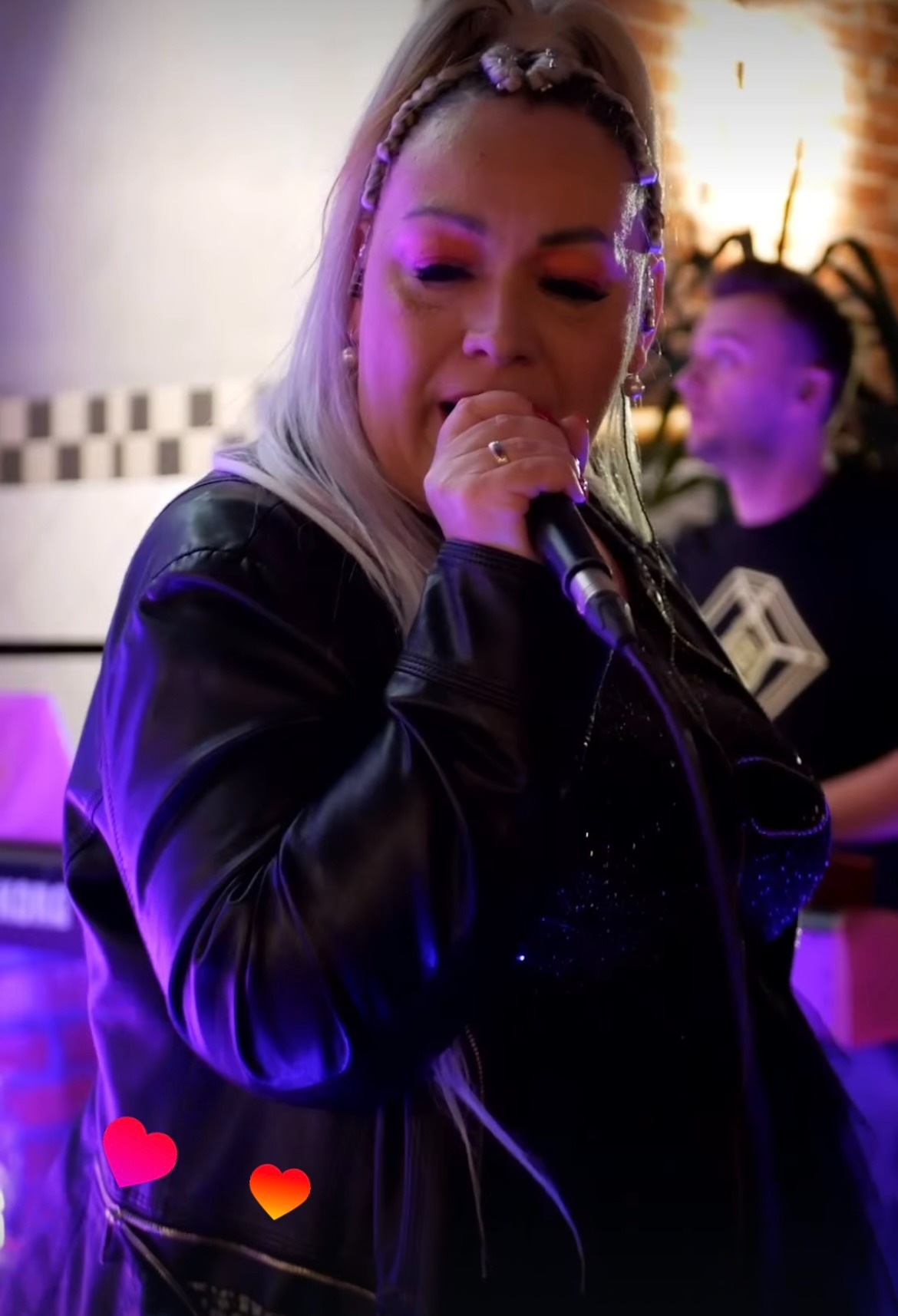 Wokalistka z mikrofonem, w skórzanej kurtce, ze srebrnymi włosami, śpiewa na scenie oświetlonej fioletowym światłem. W tle drugi muzyk.