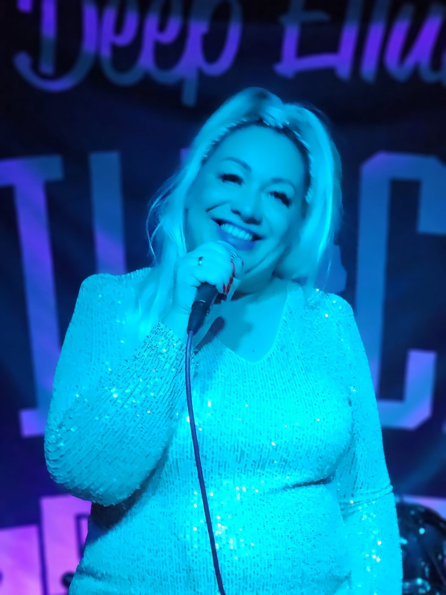 Uśmiechnięta blond wokalistka w cekinowej sukience śpiewa do mikrofonu na scenie w niebieskim oświetleniu.