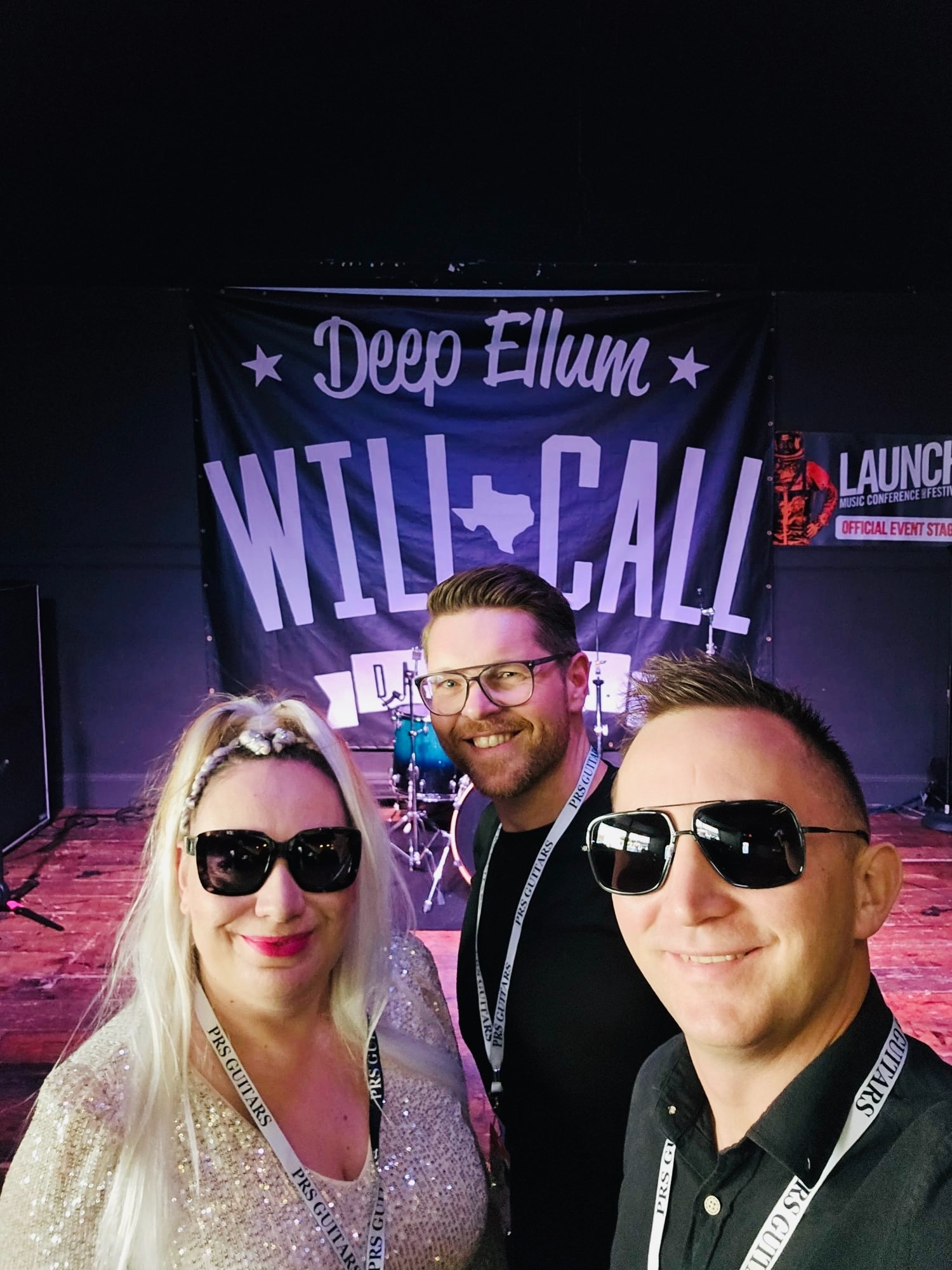 Zespół muzyczny pozuje do selfie na scenie z bębnami i banerem 'Deep Ellum Will Call' w tle. Uśmiechnięci, w okularach przeciwsłonecznych i identyfikatorach PRS Guitars.