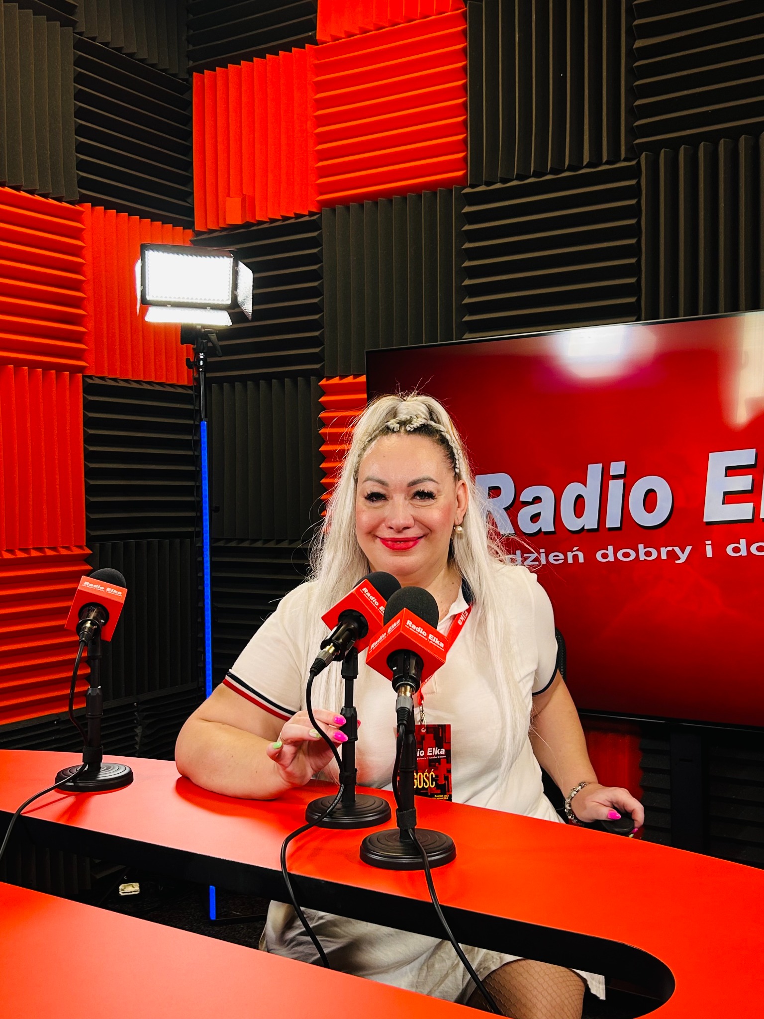 Kobieta z mikrofonami w studiu radiowym Radio Elka, z czerwonymi ścianami akustycznymi i oświetleniem LED. Uśmiechnięta prezenterka z warkoczykami.