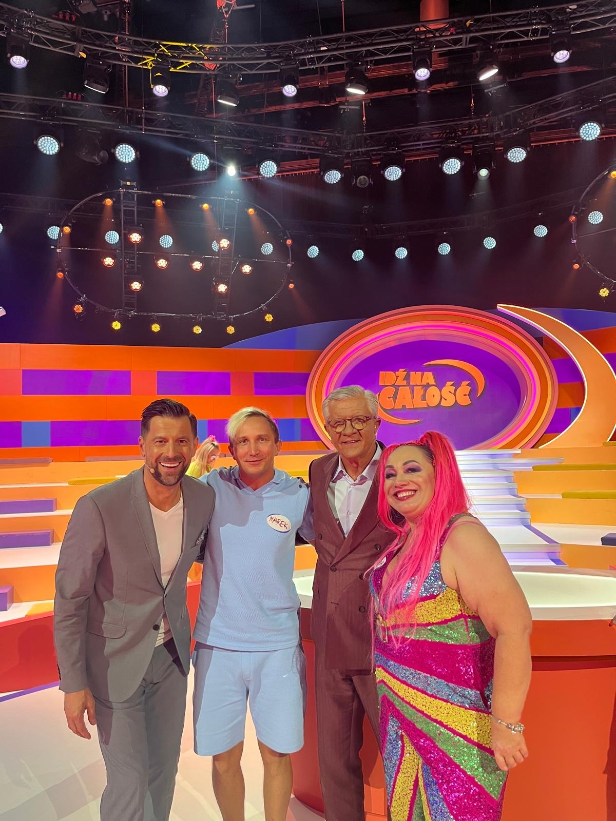 Cztery osoby (w tym piosenkarka z różowymi włosami) pozują na scenie programu telewizyjnego „Dź na całość” z oświetleniem i dekoracjami. Uśmiechnięci.
