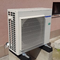 ECO POMPY MARIUSZ DOBROWOLSKI - Zewnętrzna jednostka klimatyzacji Panasonic R32 zamontowana na betonowych wspornikach przy ścianie budynku o dwukolorowej elewacji. Widoczne detale montażowe.