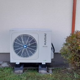 ECO POMPY MARIUSZ DOBROWOLSKI - Zewnętrzna jednostka pompy ciepła Kaisai zamontowana przy białej ścianie budynku, obok krzewu w jesiennych barwach, na trawniku.