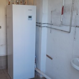 ECO POMPY MARIUSZ DOBROWOLSKI - Biała pompa ciepła Kaisai w pomieszczeniu z instalacją hydrauliczną. Widoczne rury, zbiornik wyrównawczy i płytki na podłodze. Pomieszczenie w trakcie użytkowania, ściany wymagają odświeżenia.