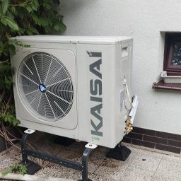 ECO POMPY MARIUSZ DOBROWOLSKI - Zewnętrzna jednostka klimatyzacji Kaisai na metalowym stelażu, obok ściany z oknem i pnącą roślinnością, na tle jasnej elewacji i kostki brukowej.