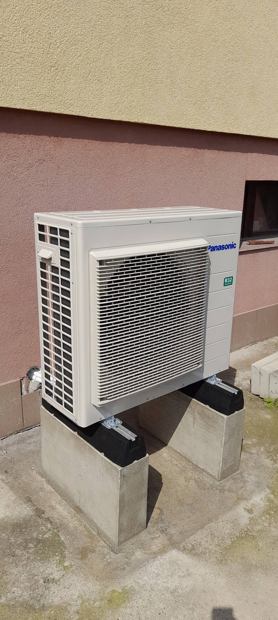 Zewnętrzna jednostka klimatyzacji Panasonic R32 zamontowana na betonowych wspornikach przy ścianie budynku o dwukolorowej elewacji. Widoczne detale montażowe.