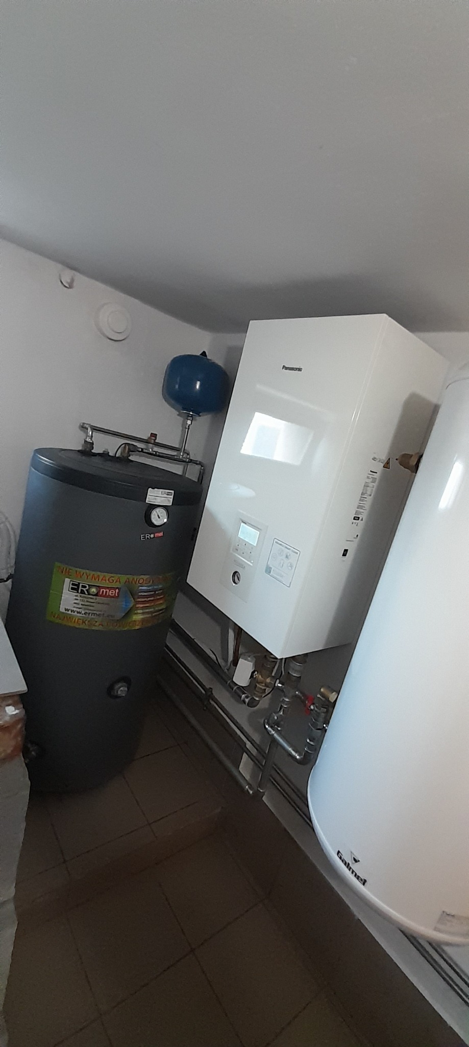 Biała pompa ciepła Panasonic i szary zbiornik ERmet z niebieskim naczyniem wzbiorczym, instalacja w Nysie. Widoczne rury i elementy instalacji.