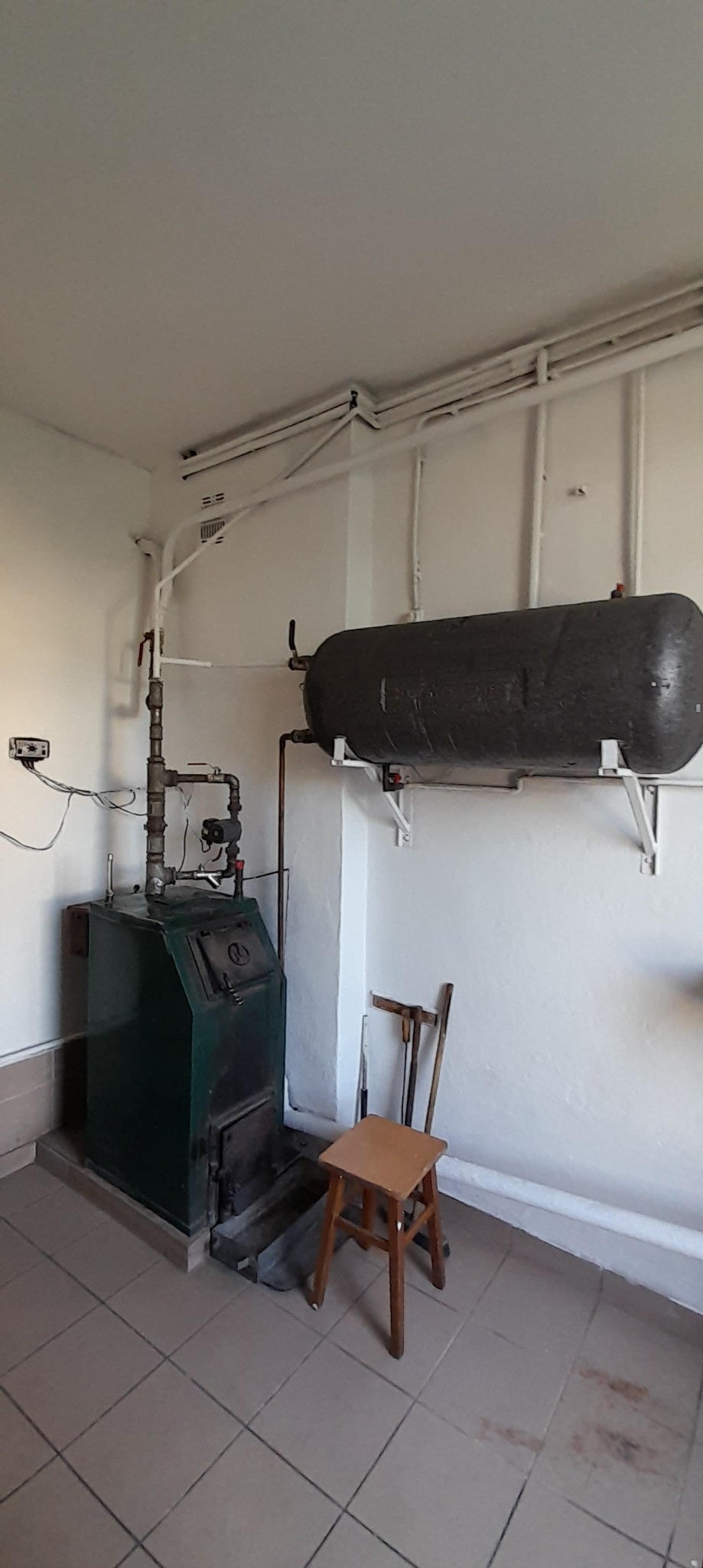 Kocioł grzewczy z instalacją i bojlerem w pomieszczeniu kotłowni, obok stoi drewniany taboret. Widoczne rury instalacji grzewczej.