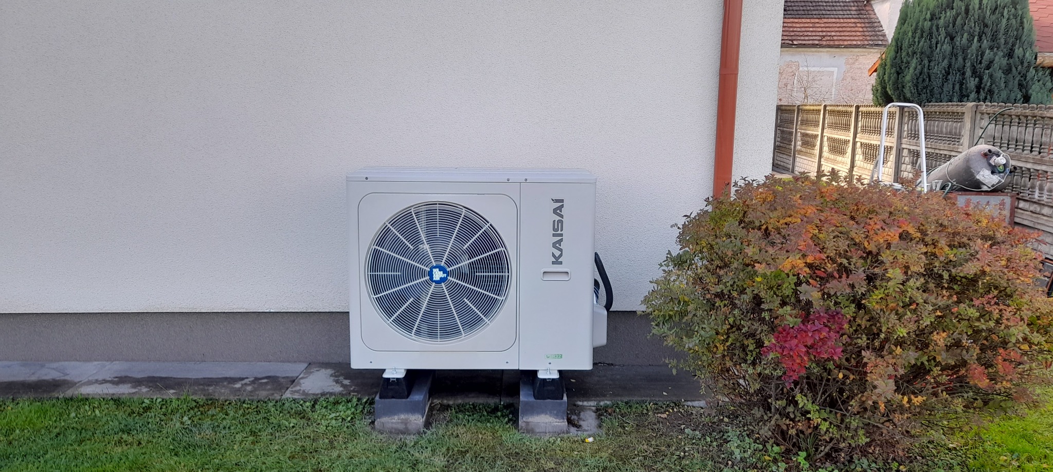 Zewnętrzna jednostka pompy ciepła Kaisai zamontowana przy białej ścianie budynku, obok krzewu w jesiennych barwach, na trawniku.