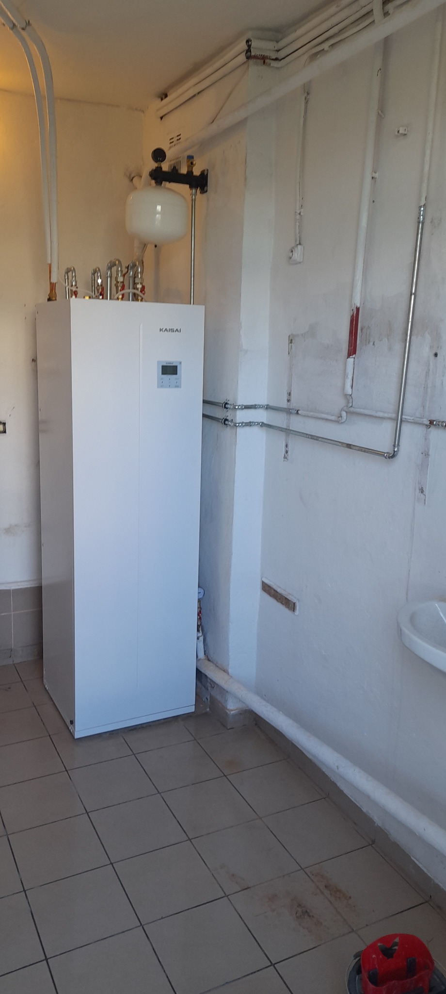 Biała pompa ciepła Kaisai w pomieszczeniu z instalacją hydrauliczną. Widoczne rury, zbiornik wyrównawczy i płytki na podłodze. Pomieszczenie w trakcie użytkowania, ściany wymagają odświeżenia.