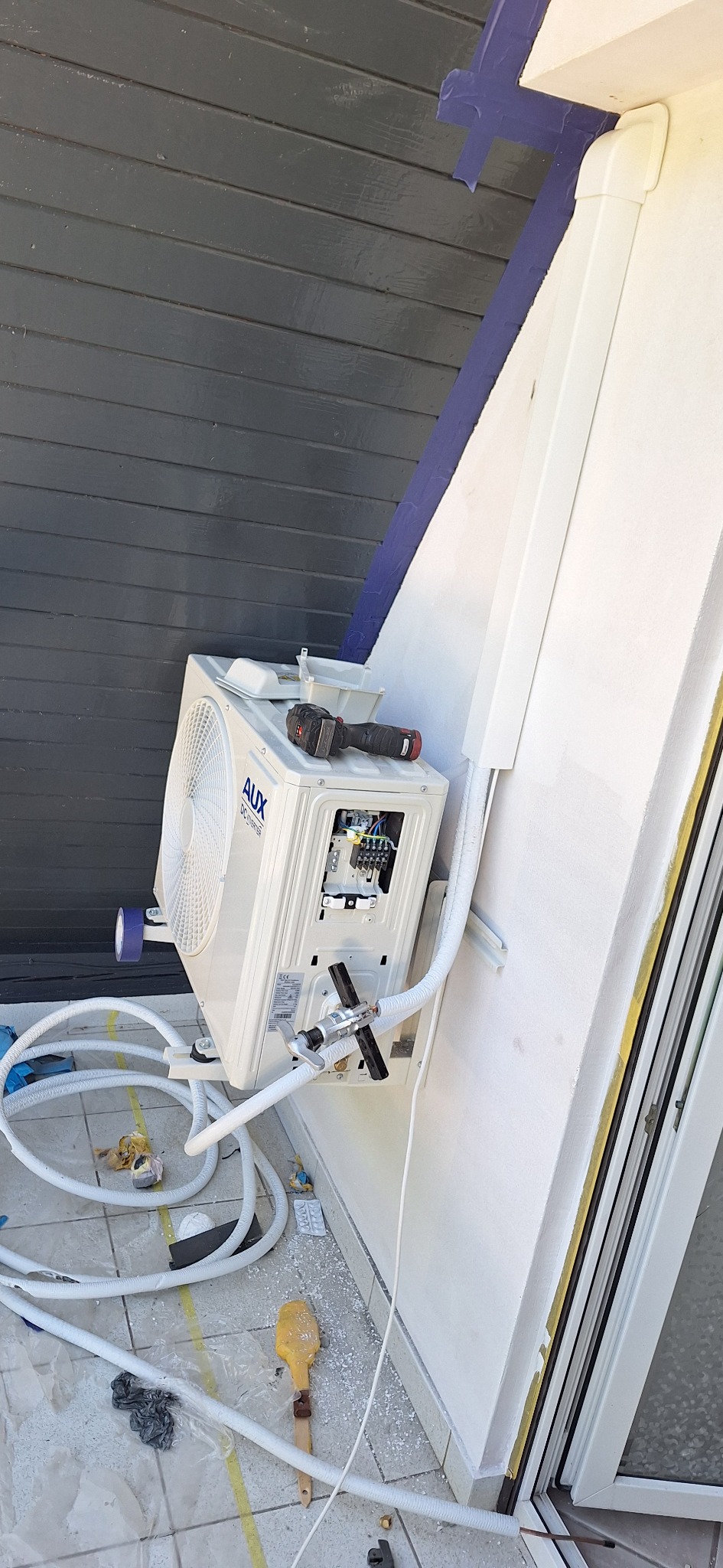 Montaż klimatyzatora AUX DC Inverter na zewnątrz budynku. Widoczne przewody, narzędzia i elementy instalacji. Ściana pomalowana na biało, szara elewacja w tle.