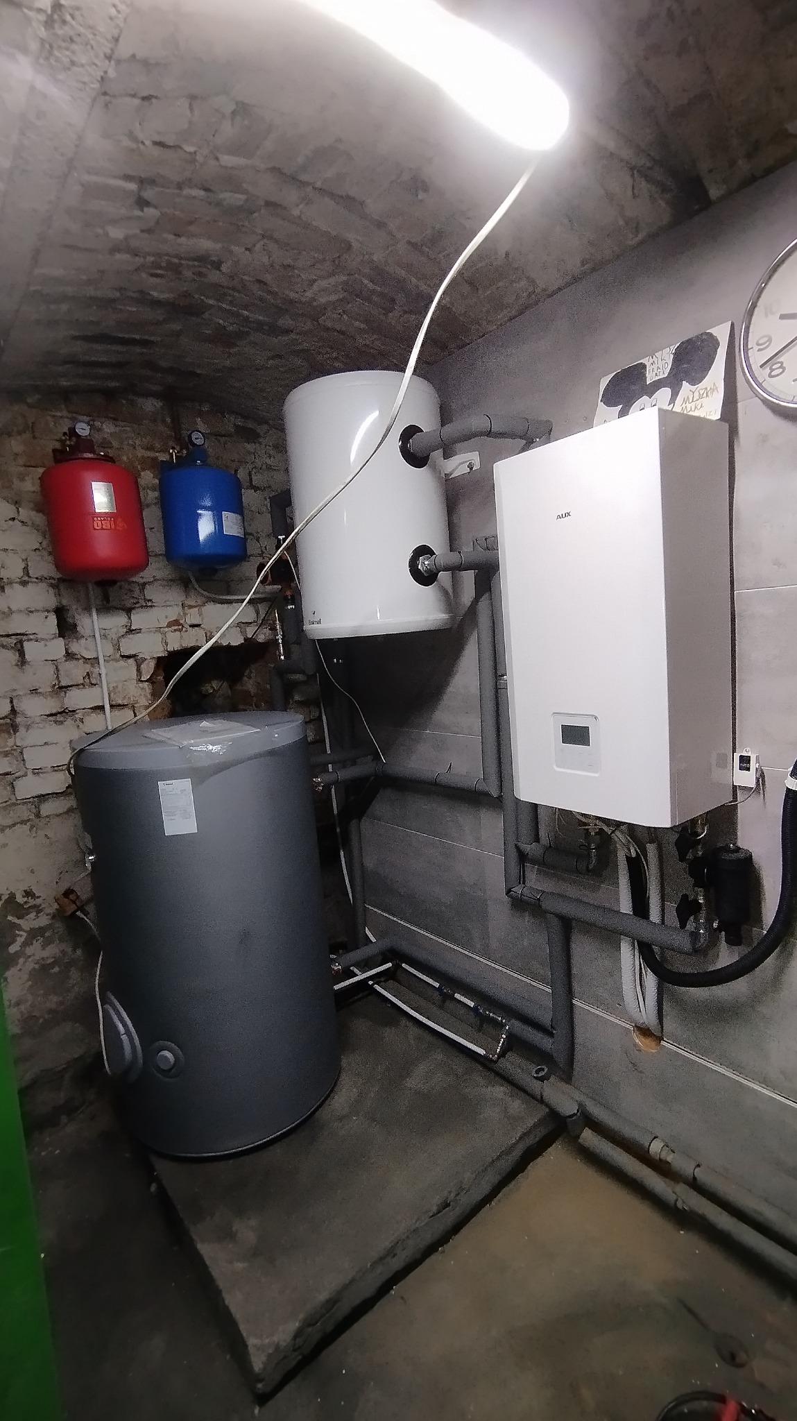 Instalacja grzewcza w piwnicy: biały bojler, szary zbiornik akumulacyjny, rury, czerwony i niebieski osprzęt na ceglanej ścianie, oświetlenie jarzeniowe.