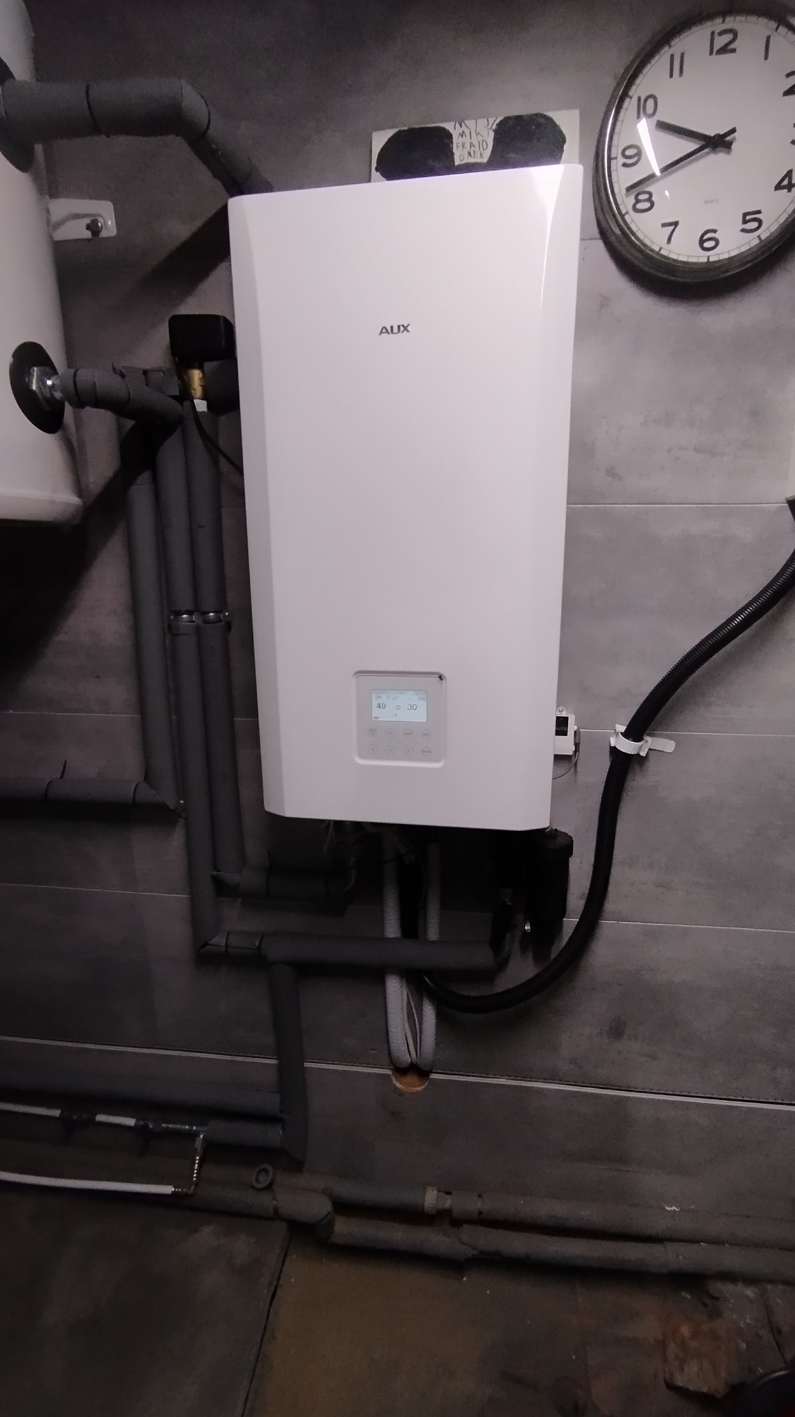 Biała pompa ciepła AUX zamontowana na szarej ścianie z widocznymi rurami instalacyjnymi i zegarem. Wyświetlacz pompy wskazuje temperaturę.