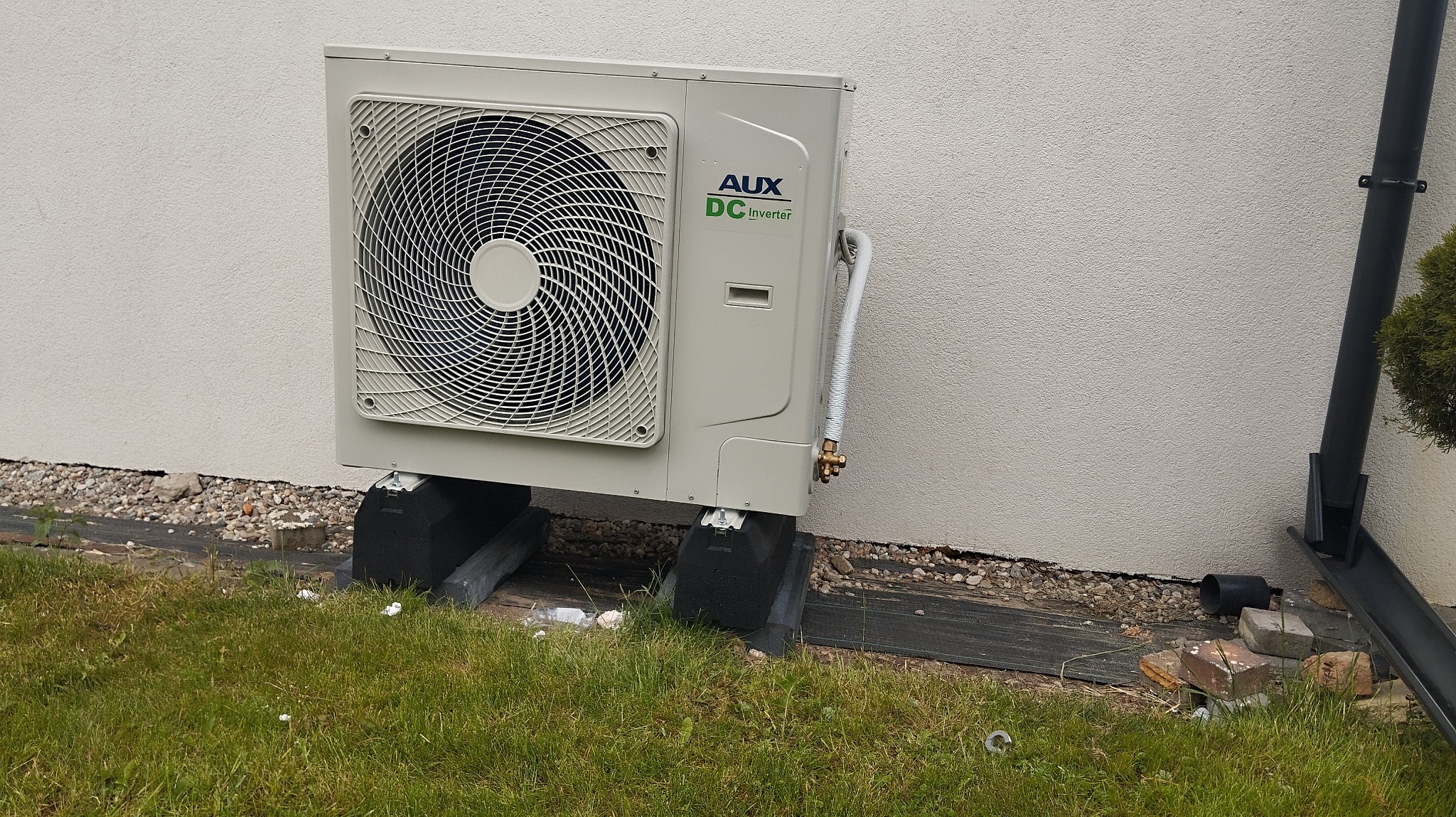 Szara pompa ciepła AUX DC Inverter, zamontowana na czarnych podkładkach przy ścianie budynku, z widoczną rurą odprowadzającą. Trawa i kamyki w tle.