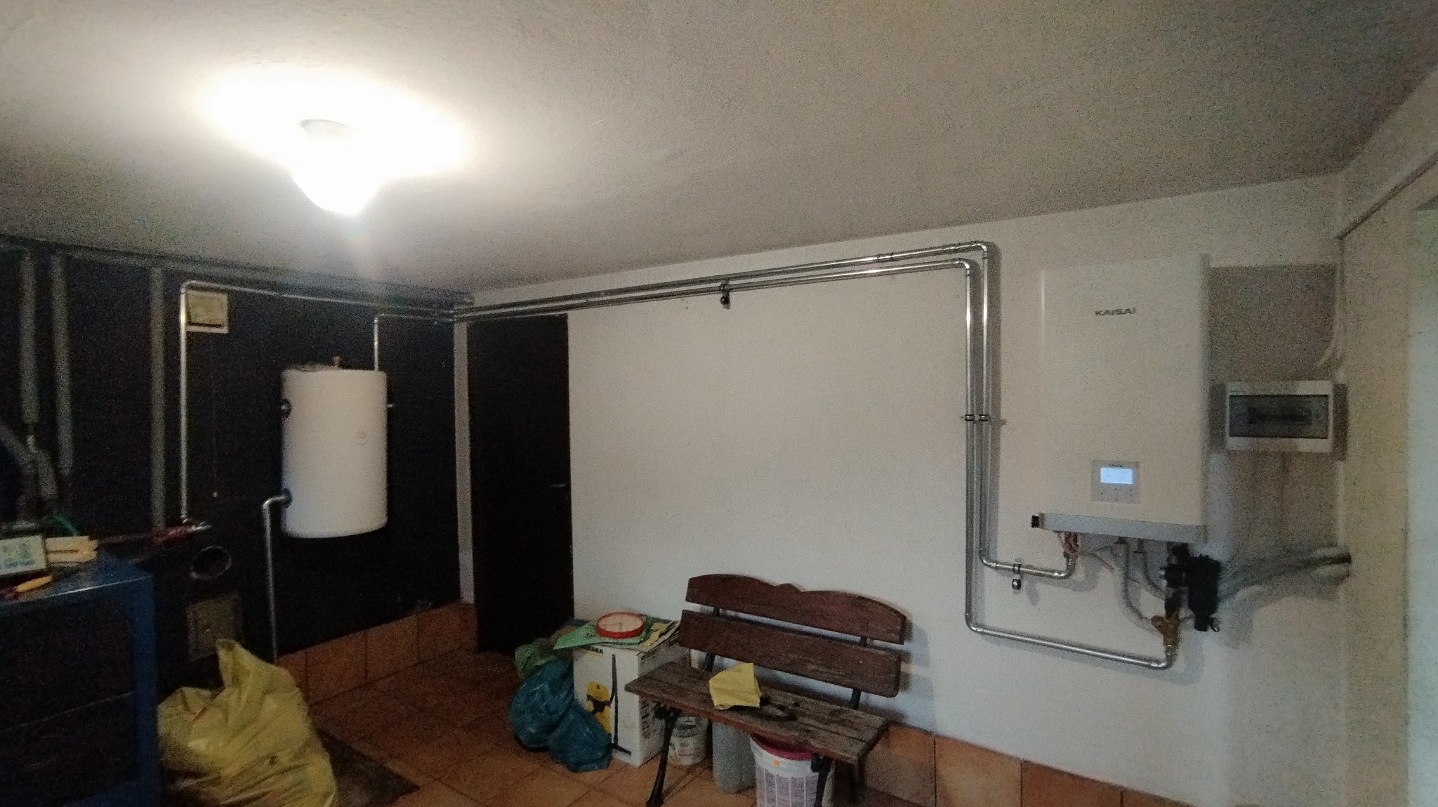 Kotłownia z zamontowanym białym bojlerem i pompą ciepła Kaisai. Widoczne rury instalacyjne, drewniana ławka i narzędzia.